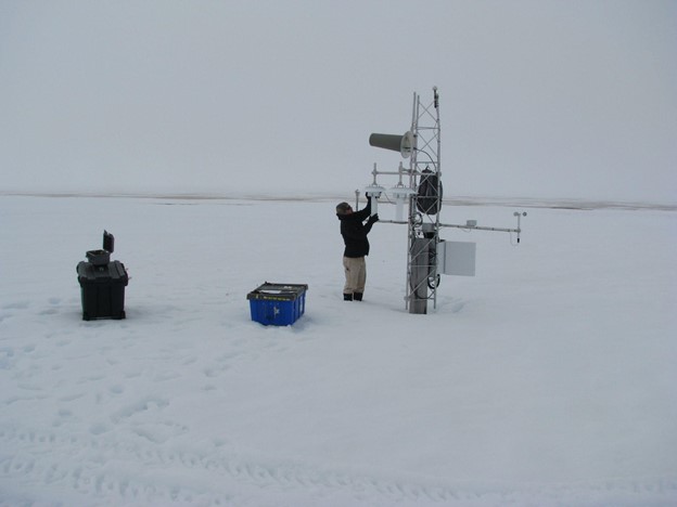 Snow covered field with a man working on a short tower. Boxes are scattered around. 