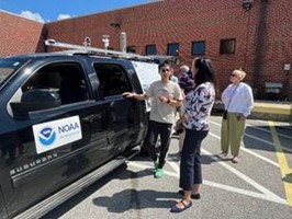 Three people standing around a black SUV with the NOAA logo on the side. Various instruments are mounted to the roof.