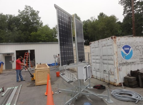 man standing next to a large wood crate. Two solar panels, and a shipping container with the NOAA logo appear in the photo. There are boxes, tools, cables and pipes scattered around.