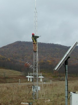 man wearing a hard hat climbing tall metal tower in a field with a mountain in the distance