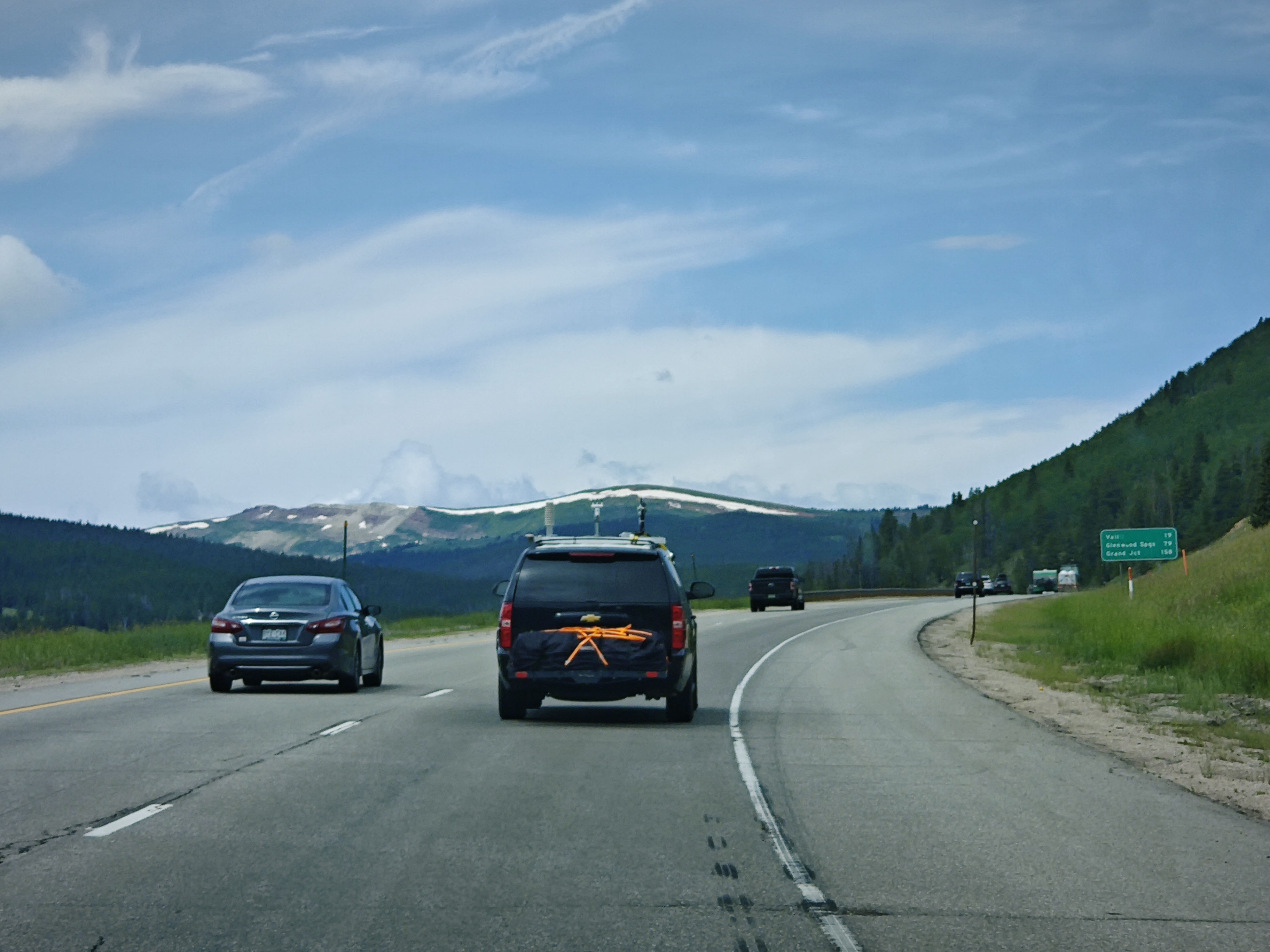 photo taken from a car on a highway with an SUV and a sedan ahead. Expansive blue sky and some rolling hills ahead on the road