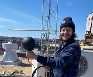 smiling man holding a round, black temperature sensor in front of metal tower on top of a flat roof.