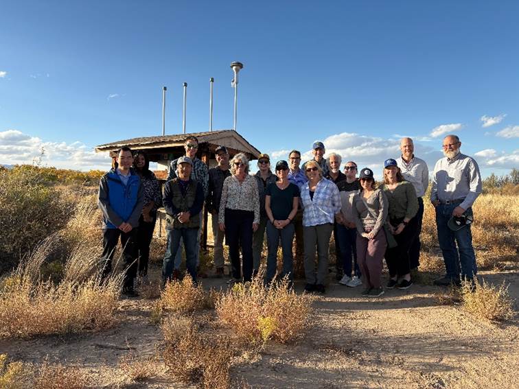 Group of people standing out in a field of yellow grasses. There is a small shelter behind them with sensors mounted on top.